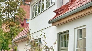 Une maison moderne à deux étages, avec un toit recouvert de tuiles rouges, des murs extérieurs blancs et un petit jardin à l’avant, agrémenté de diverses plantes et d’un banc.