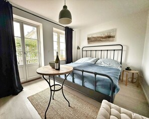 A bedroom with a bed, a round table with a bottle, a small lamp, and a window with curtains. - Romantic apartment for 2 people - 200m to the Strelasund with a view of Stralsund (Altefähr)