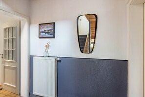 A hallway with a white door, a framed picture of a cityscape, a radiator, and a mirror reflecting a staircase.