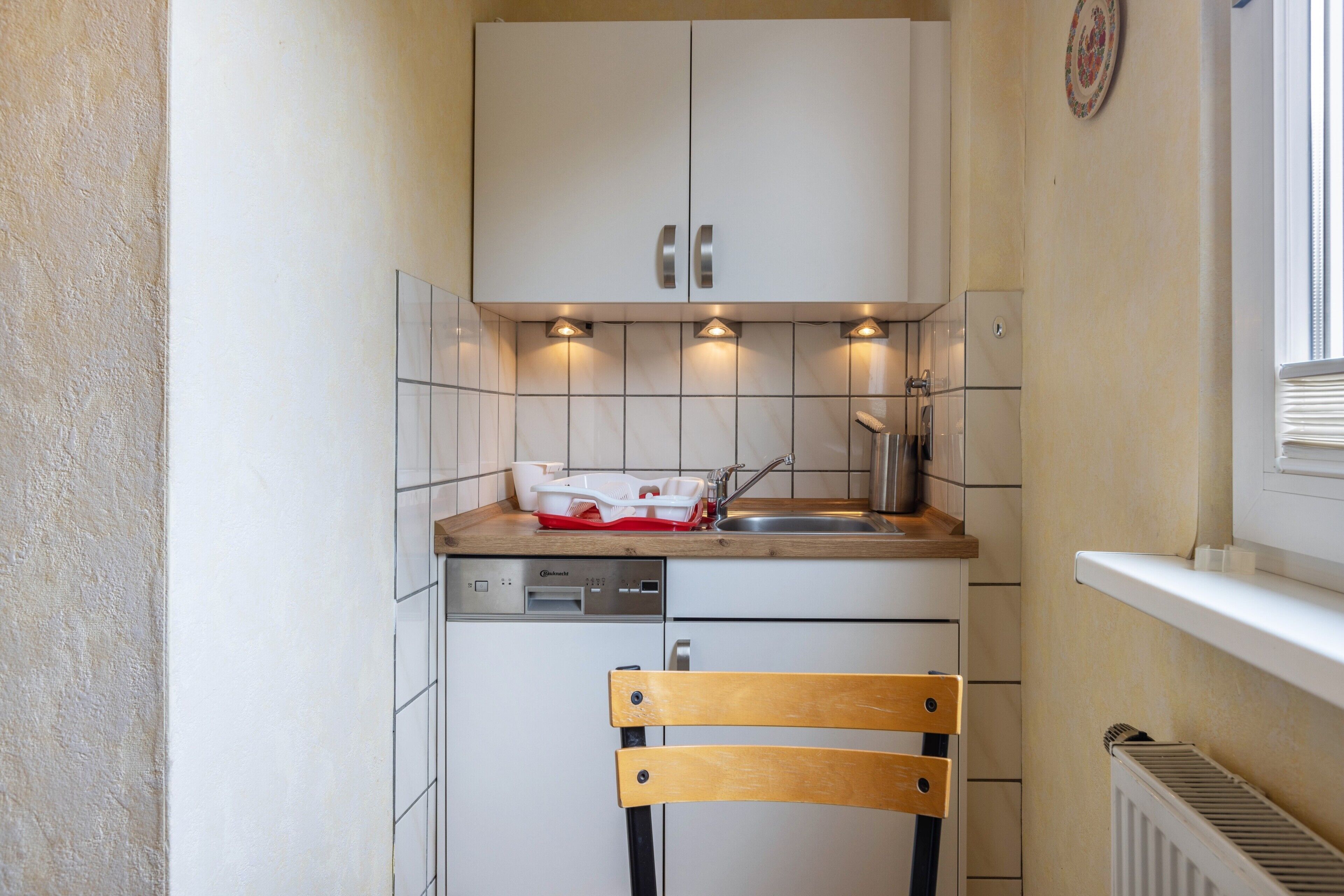 A compact kitchen with white cabinets, a built-in dishwasher, a wooden chair, and a window with blinds.