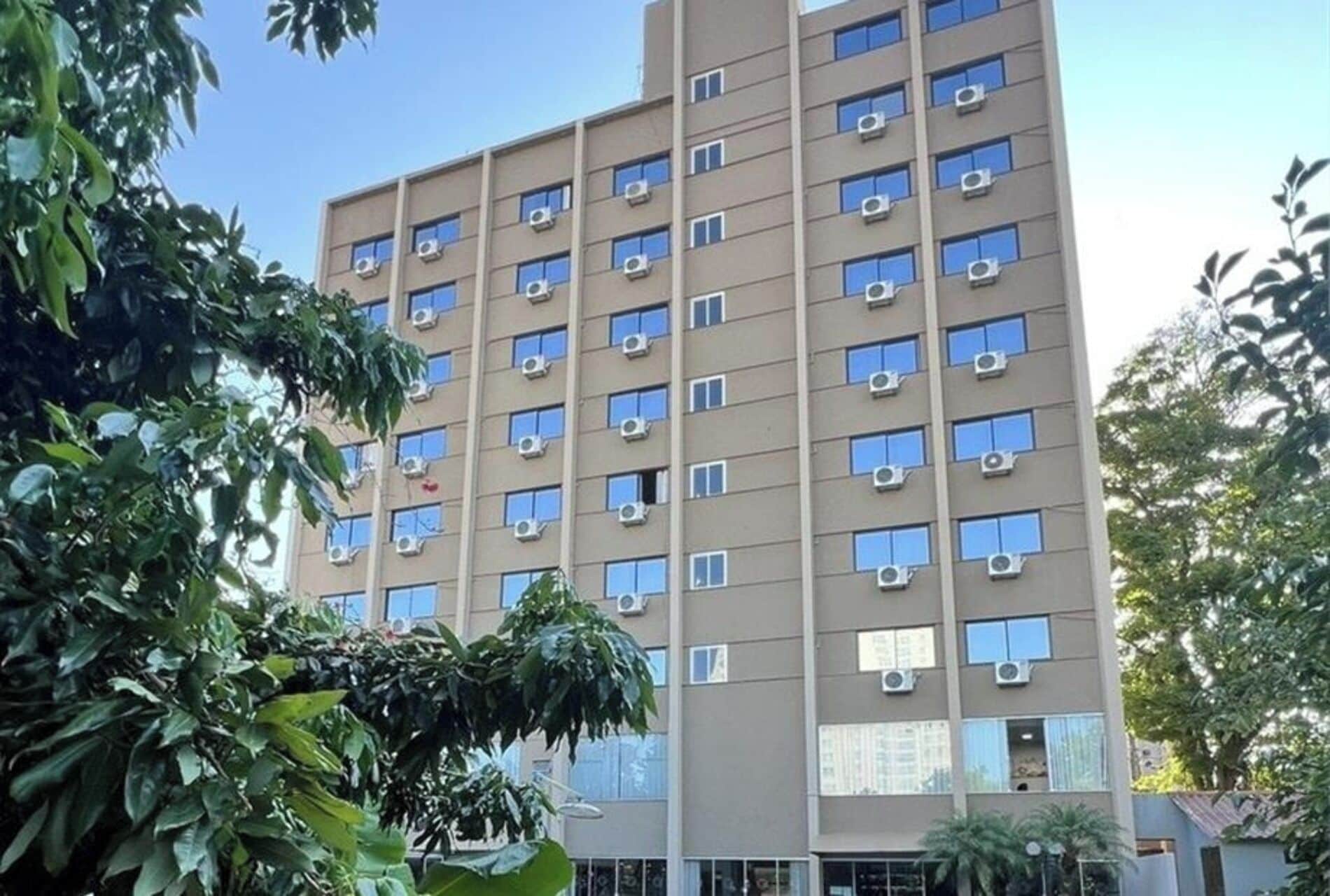 Un edificio de varios pisos con numerosas ventanas y unidades de aire acondicionado, rodeado de vegetación.