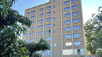 Un edificio de varios pisos con numerosas ventanas y unidades de aire acondicionado, rodeado de vegetación.