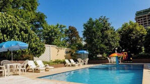 A swimming pool with lounge chairs and umbrellas, surrounded by trees and a building in the background.