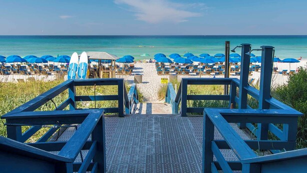 Blick auf einen Strand mit einer Holztreppe, die zu Liegestühlen und Sonnenschirmen führt.