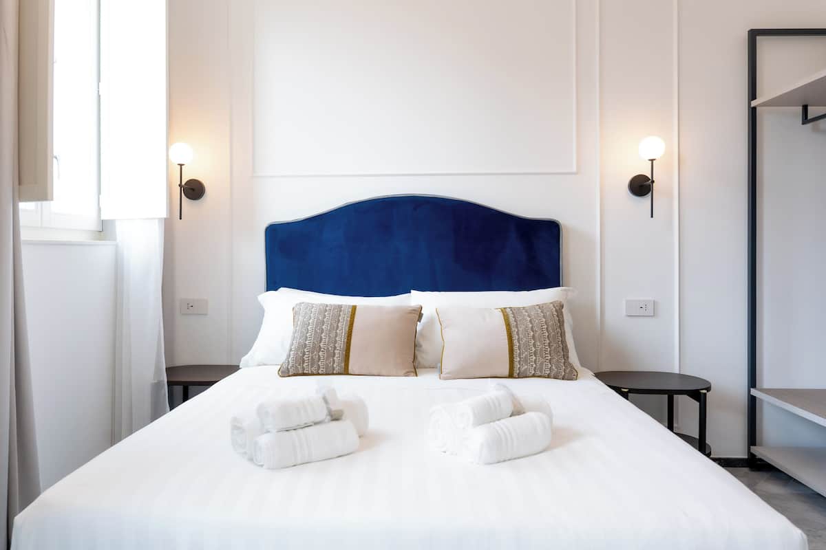Un lit bien fait, avec du linge de lit blanc et deux serviettes enroulées, une tête de lit bleue et deux tables de chevet équipées de lampes fixées au mur.