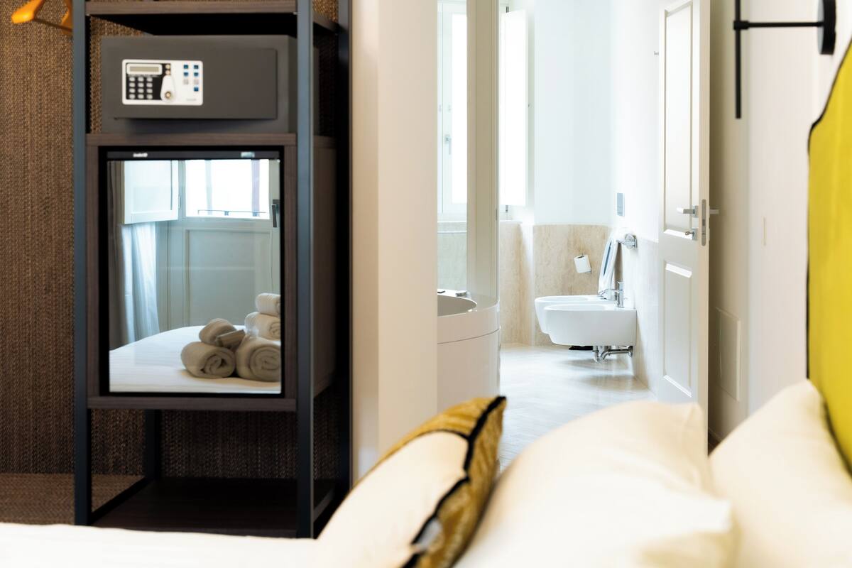 Une chambre d’hôtel avec un lit, une table de chevet dotée d’un panneau de commande, une vue sur la salle de bain et un oreiller jaune.