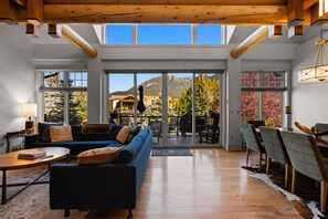 Amplia sala de estar con un sofá azul, una mesa de centro de madera y un comedor con sillas y mesa. Grandes ventanales ofrecen vistas a montañas y árboles.