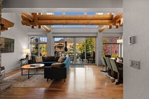A spacious living room with a wooden ceiling, large windows, a sofa, a coffee table, and a dining area with chairs.