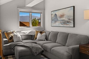A modern living room with a grey sectional sofa, a window with a view of mountains, and a painting on the wall.