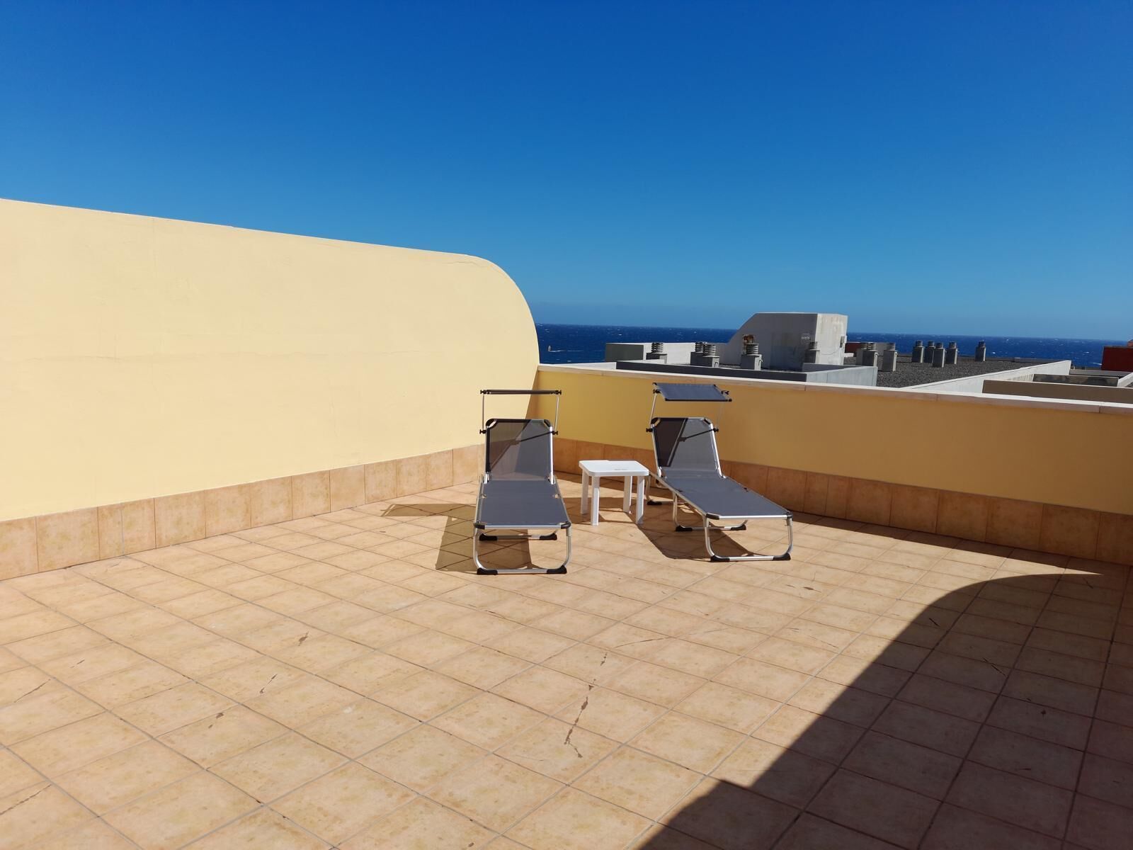 A rooftop terrace with two sun loungers and a small table, overlooking a clear blue sky and the sea.