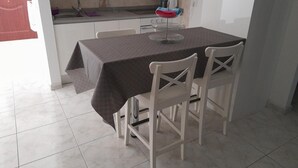 Ein Essbereich mit Tisch, bedeckt mit einem dunklen Tuch, weißen Stühlen, einer Küche im Hintergrund und einem gefliesten Boden.