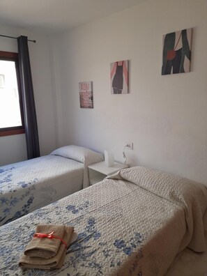 Een kamer met twee bedden, een klein tafeltje en een raam met gordijnen. Er hangen drie kunstwerken aan de muur.