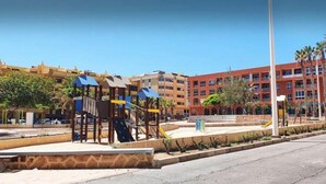 Ein Spielplatz mit Rutschen und Klettergerüsten, umgeben von Gebäuden mit Balkonen und Palmen.
