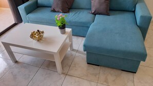 Ein smaragdgrünes Ecksofa mit zwei Kissen, ein weißer Couchtisch mit einer dekorativen Schale und einem Topf mit Pflanze und ein gefliester Boden.