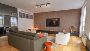 A modern living room with a sectional sofa, a white armchair, and a coffee table. There is a flat-screen TV mounted on the wall, and the room has wooden flooring.