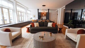 A modern living room with a dark sofa, two white armchairs, a striped coffee table, and a kitchen in the background.