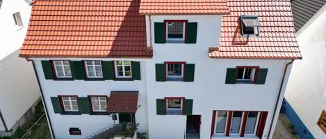 A two-story building with a red-tiled roof, multiple windows with green shutters, and a small staircase leading to the entrance.