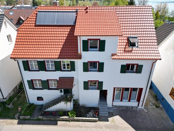 A two-story building with a red-tiled roof, multiple windows with green shutters, and a small staircase leading to the entrance.