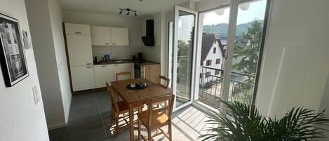 A modern apartment with a dining area, kitchen, and large sliding glass doors leading to a balcony with a view of houses.