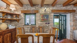 A rustic dining area with a stone wall, wooden beams, a table with chairs, a vase, and a painting.