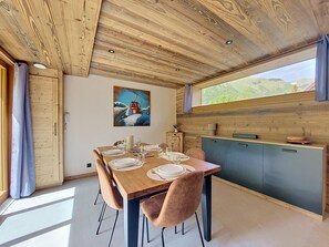 A dining area with a wooden table set for four, a painting on the wall, and a view of greenery outside.