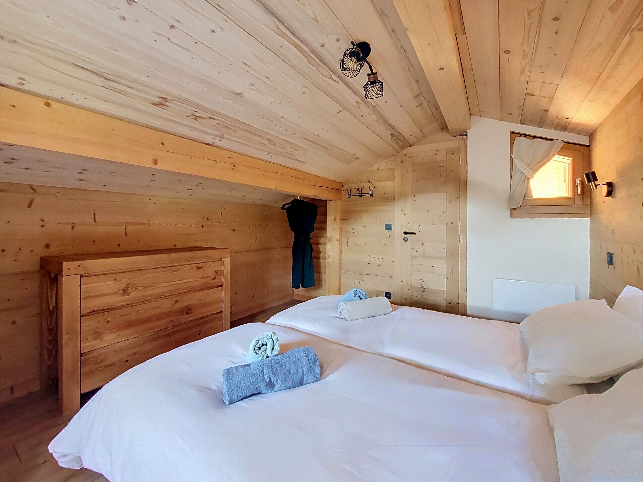 Una habitación de cabaña de madera con dos camas, un cabecero de madera, un perchero y una pequeña ventana con cortina.