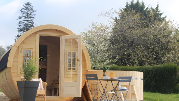 A wooden cabin with a circular design, a door open to a room, a table with chairs, and a potted plant outside. - Unique Barrel So'Happy - A Gourmet Getaway (Sevremont)