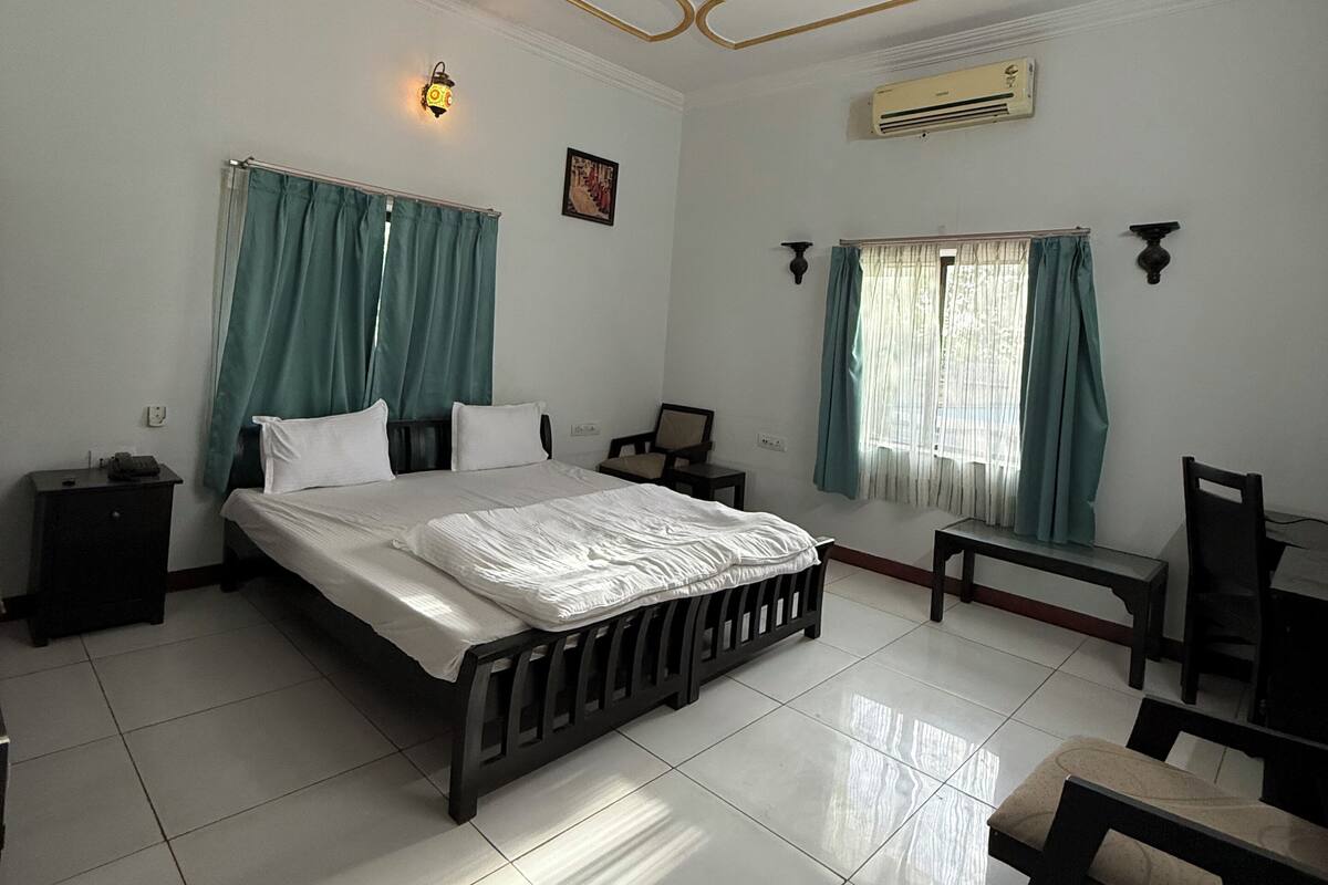 A bedroom with a large bed, a ceiling fan, an air conditioning unit, a nightstand, a chair, and a window with curtains.