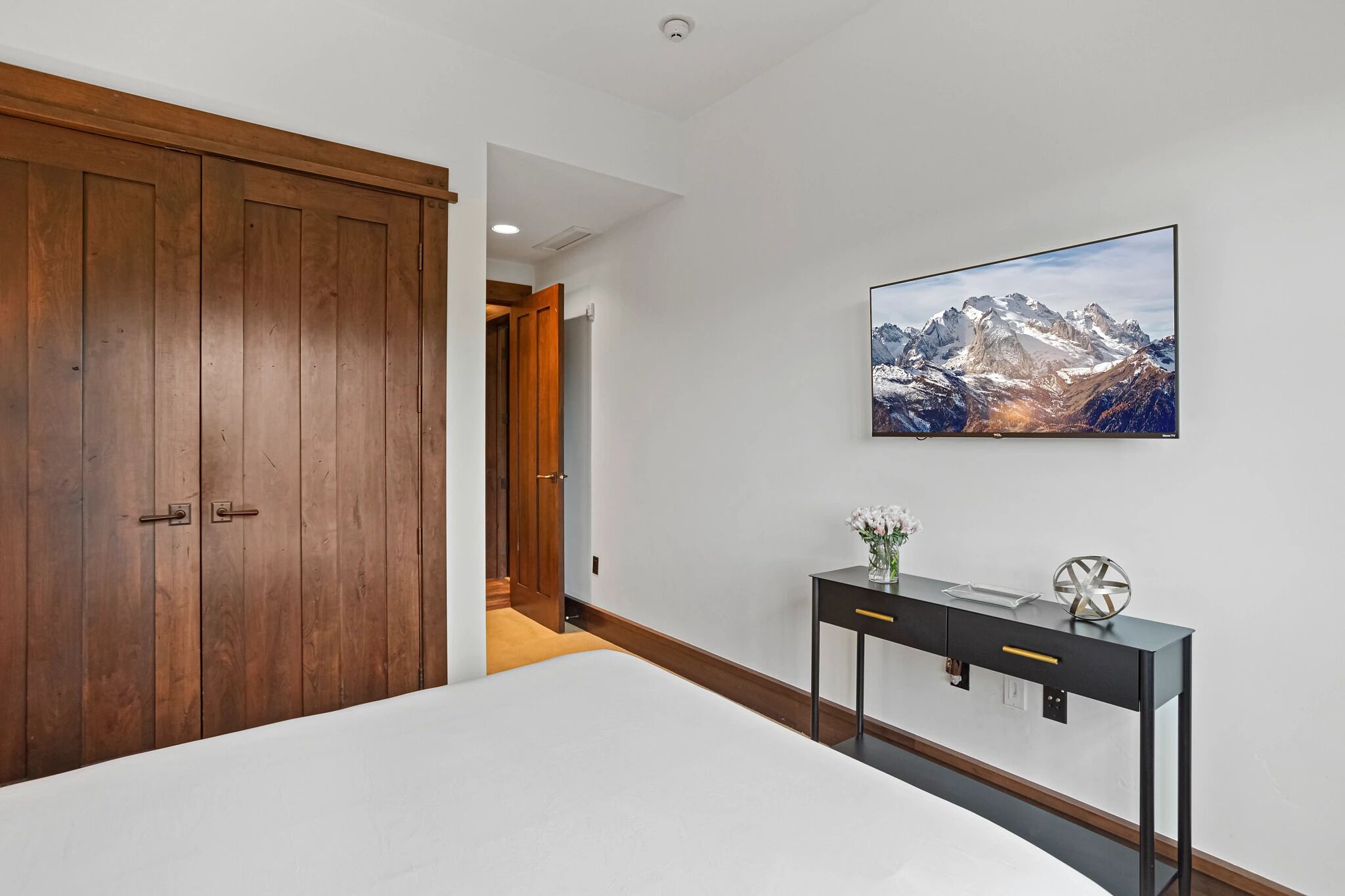Un dormitorio con una cama grande, un armario de madera, un escritorio con un jarrón y un cuadro de un paisaje montañoso en la pared.
