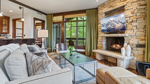 A modern living room with a stone fireplace, a large TV mounted above it, and a view of the outdoors through large windows. - Arrowleaf Lodge at Deer Valley Resort - Three Bedroom Residence #203 3 Condo (Park City)