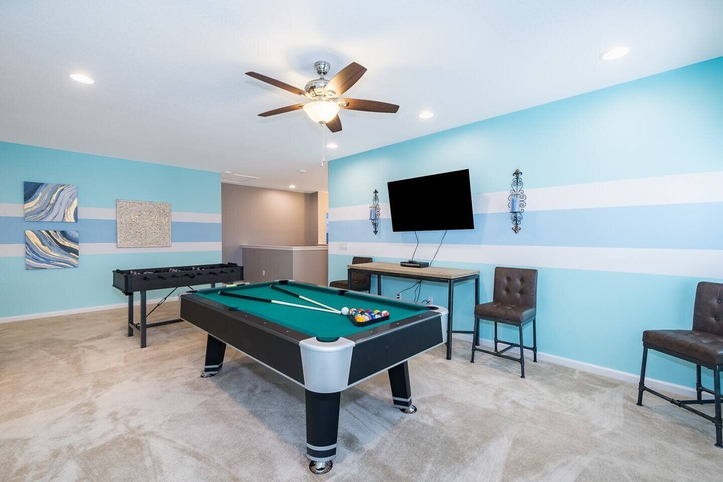 A pool table with a ceiling fan, a TV, and two chairs in a room with blue walls and carpeted flooring.
