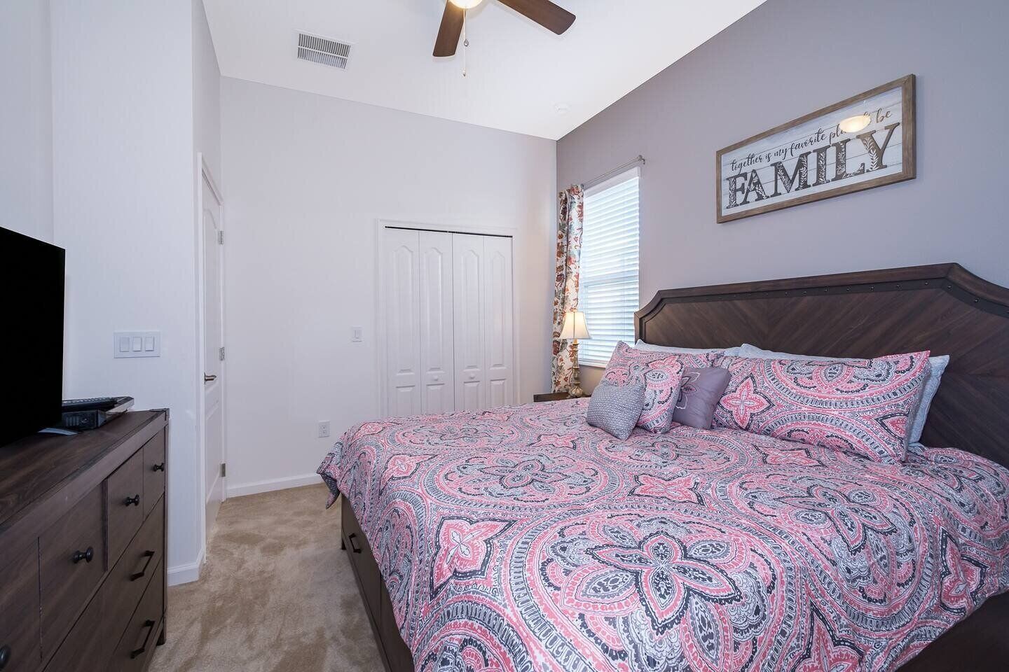 A bedroom with a bed, a dresser, a ceiling fan, a window with curtains, and a wall sign.