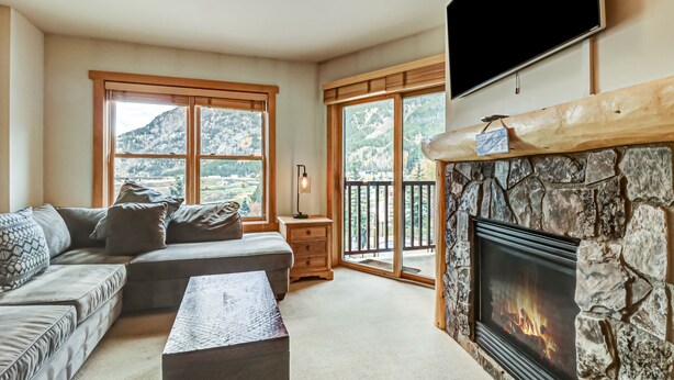 A living room with a stone fireplace, large windows with mountain views, a sofa, and a television. - Cs321 Copper Springs 1 Bedroom Condo (Copper Mountain)