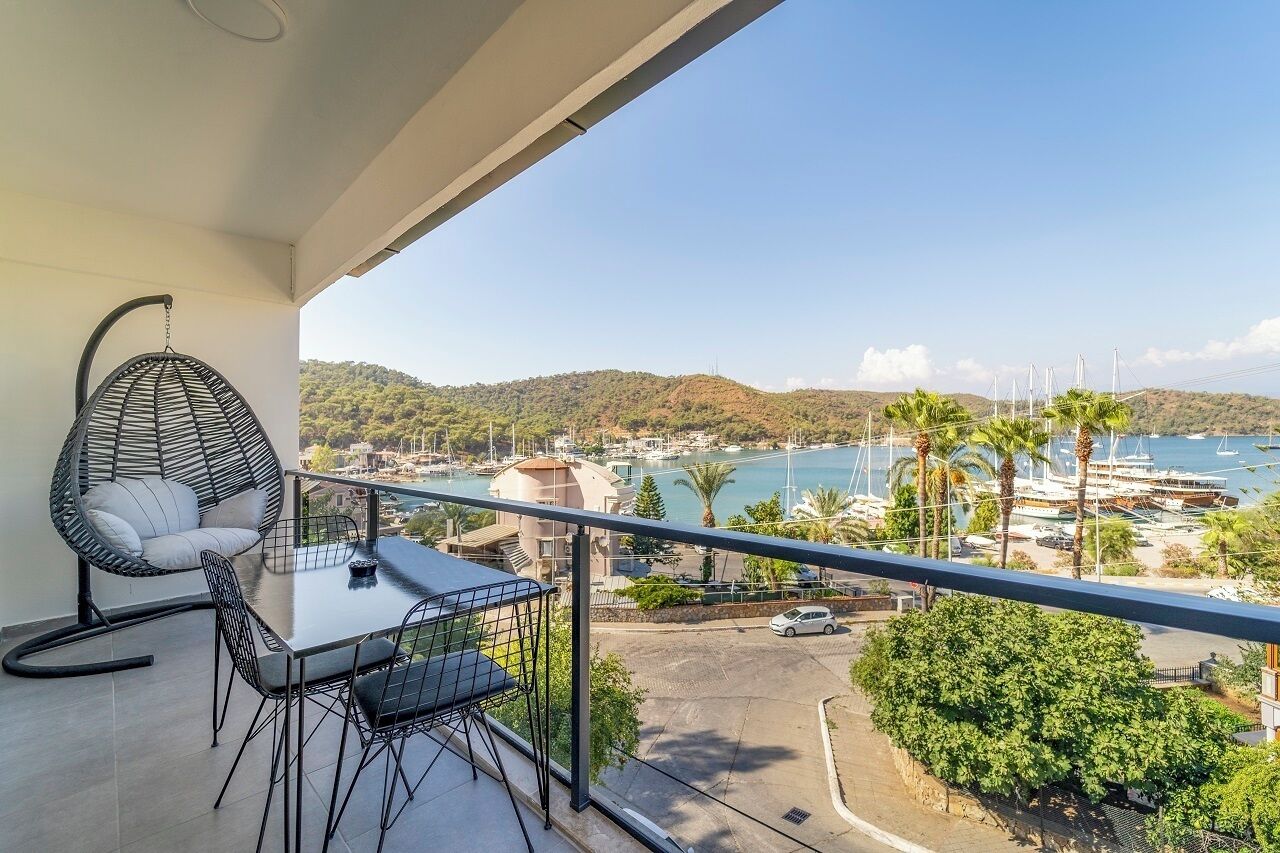 A balcony with a table and chairs, overlooking a marina with boats and a hilly landscape.