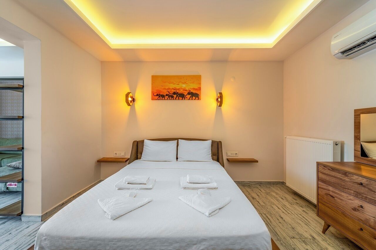 A hotel room with a bed, bedside tables, a dresser, a painting on the wall, and an air conditioning unit.