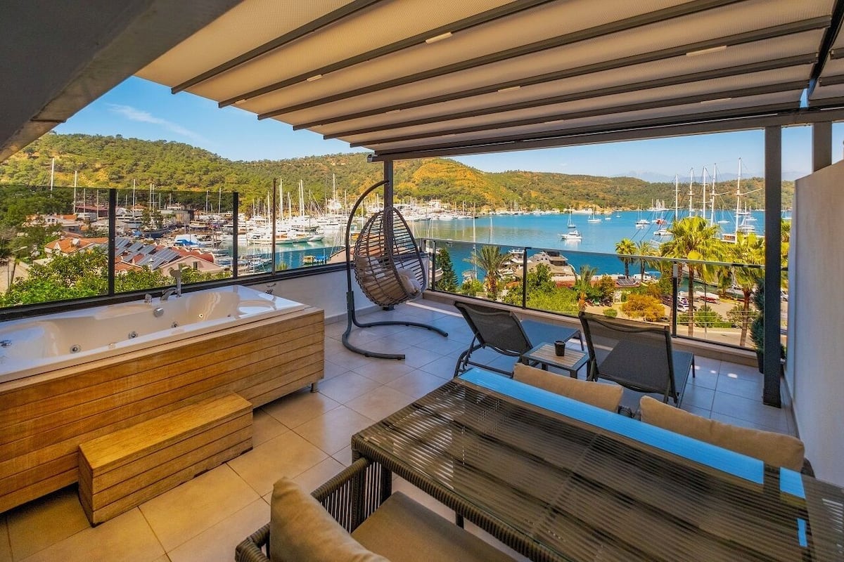 A balcony with a view of a marina, a wooden bench, a hanging chair, and a large bathtub.
