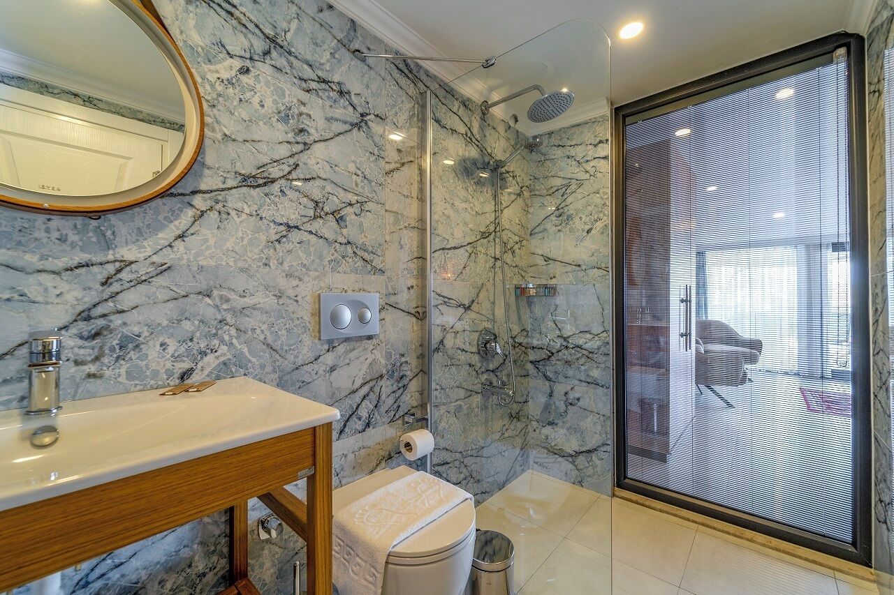 Un baño con pared de mármol, tocador de madera, cabina de ducha y un amplio ventanal.