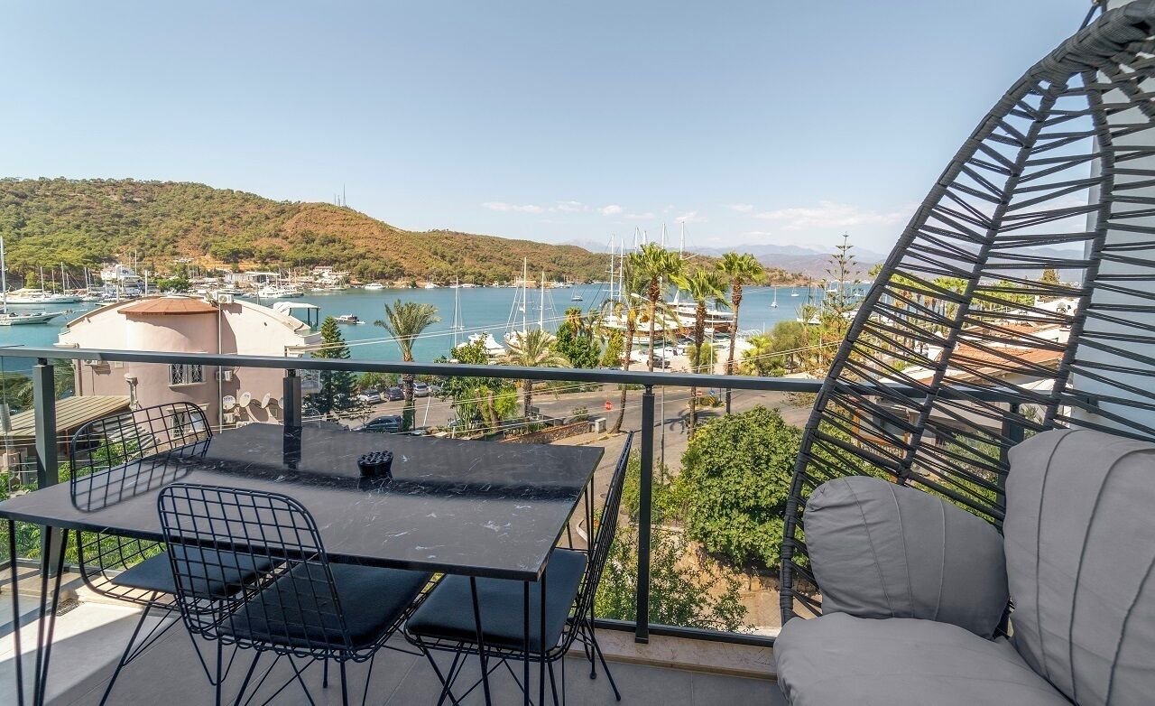 Een balkon met een tafel en stoelen, uitkijkend op een jachthaven met boten en een heuvelachtig landschap.
