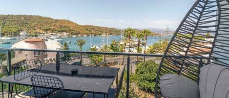 Varanda com mesa e cadeiras, vista para uma marina com barcos e uma paisagem montanhosa.