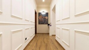 Un couloir aux murs inférieurs recouverts de lambris blancs et doté d’un plancher en bois, qui mène à une pièce où est accrochée une photographie encadrée d’un bâtiment.