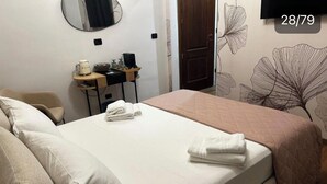 Un lit bien fait, avec des oreillers et une serviette pliée, une petite table de chevet avec une bouilloire et des articles de toilette, et un mur orné d’un motif floral.