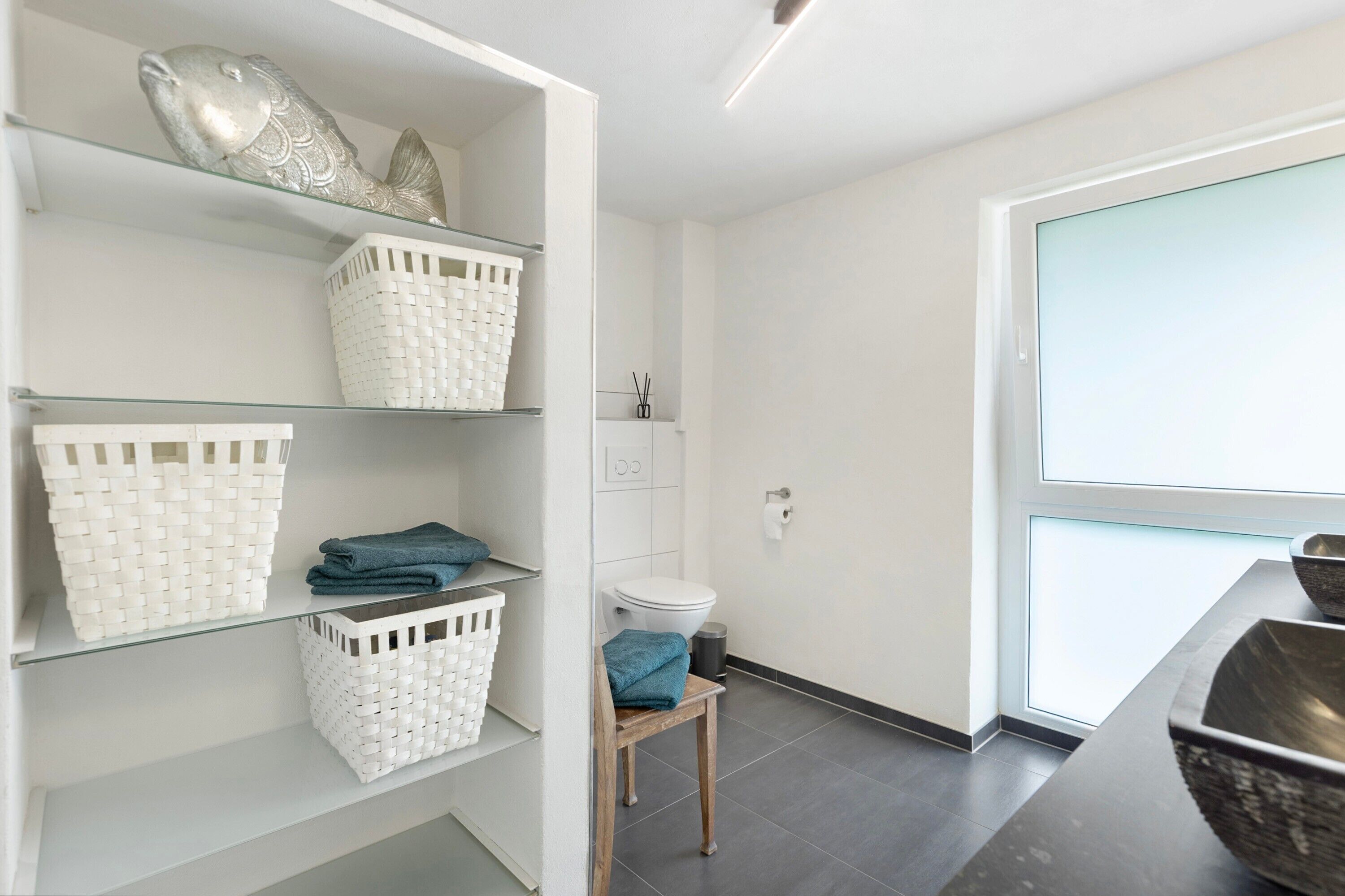 A modern bathroom with a toilet, shelves with baskets, and a window.