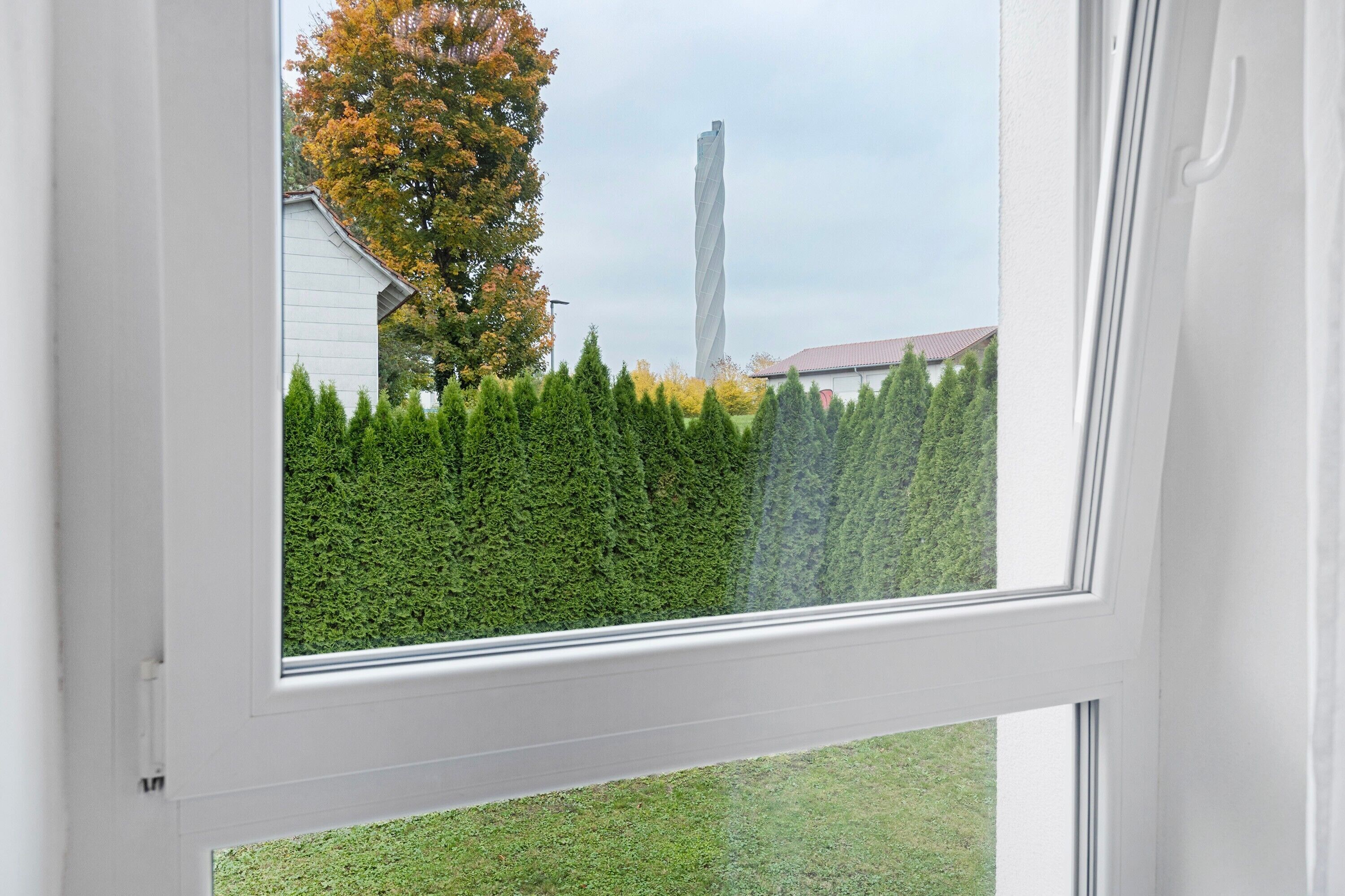 A window with a view of a white building, a twisted tower-like structure, and a hedge.