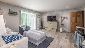 A modern living room with a sofa, ottoman, TV, wooden cabinets, and a brick wall. - Blue Wave Suites (Clearwater Beach)