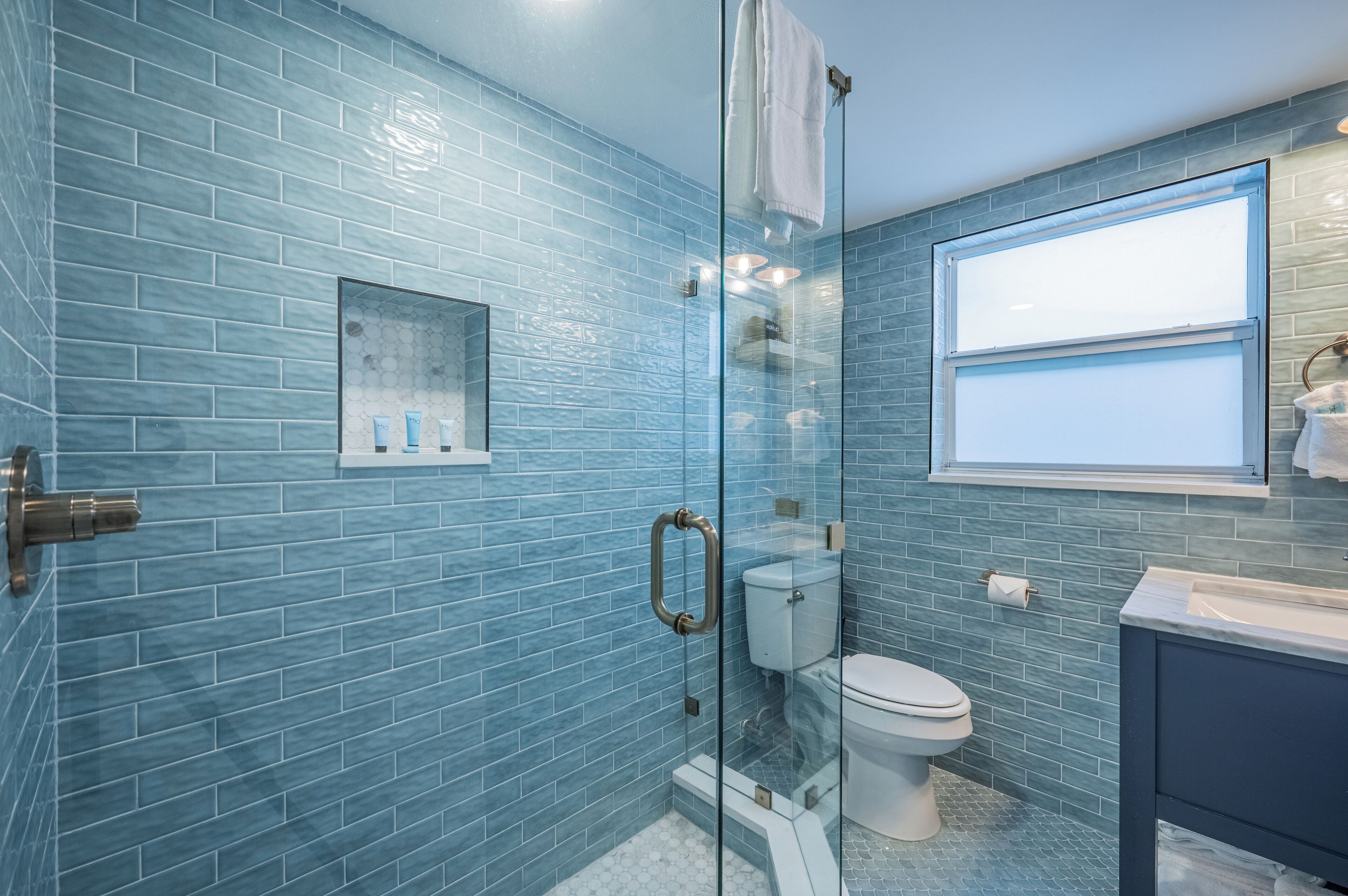 A bathroom with blue-tiled walls, a glass shower enclosure, a toilet, and a sink with a window above it.