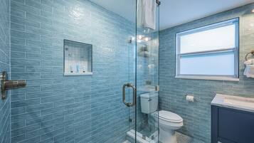 A bathroom with blue-tiled walls, a glass shower enclosure, a toilet, and a sink with a window above it.
