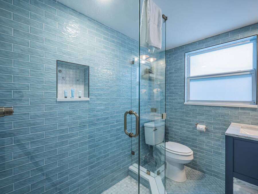 A bathroom with blue-tiled walls, a glass shower enclosure, a toilet, and a sink with a window above it.
