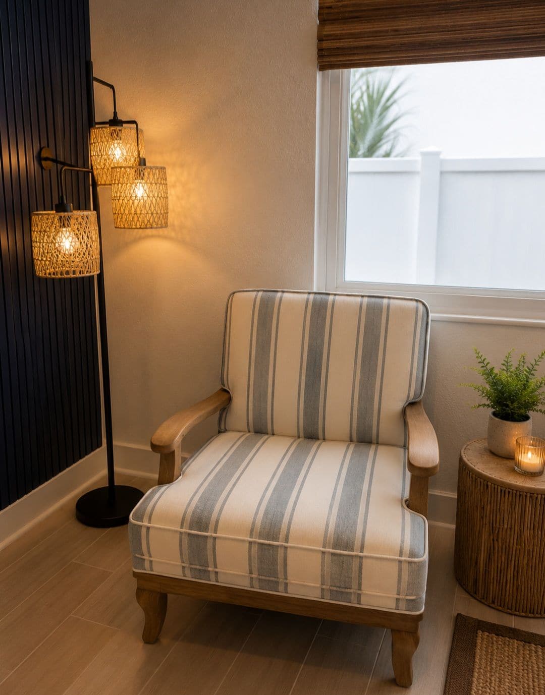 Un fauteuil à rayures avec des accoudoirs en bois, placé à côté d’un lampadaire doté d’un abat-jour en tissu tressé, et une petite table ronde sur laquelle se trouvent une plante et une bougie.