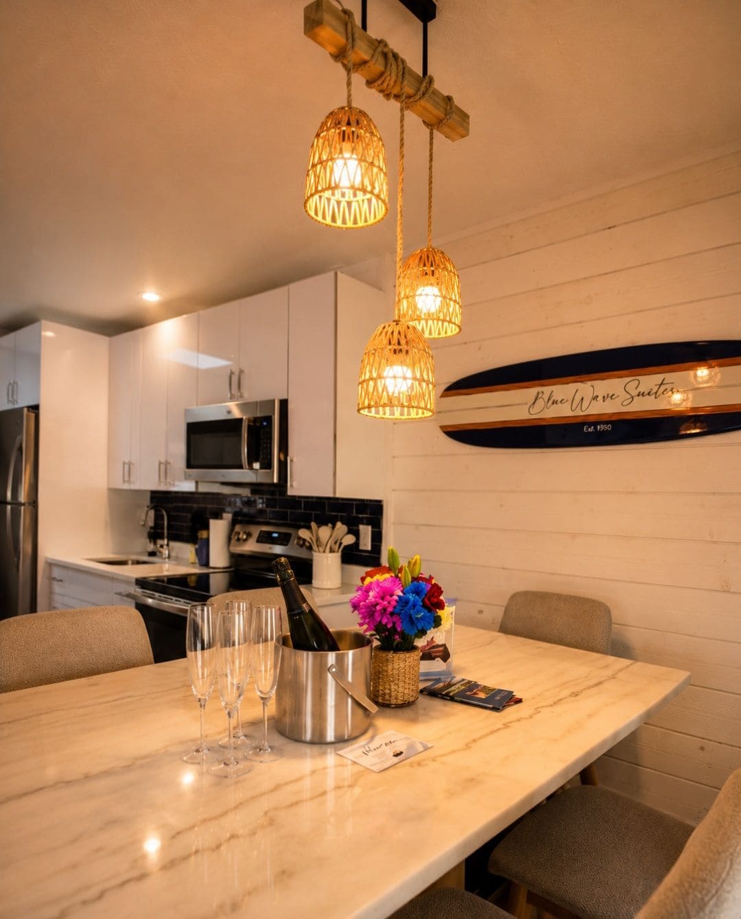 A modern kitchen with white cabinets, a stainless steel refrigerator, and a microwave. There are two hanging wicker lamps, a surfboard decoration, and a table with wine glasses and a bottle.