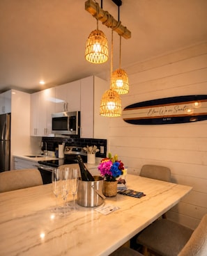 Een moderne keuken met witte kastjes, een roestvrijstalen koelkast en een magnetron. Twee hangende rieten lampen, een surfboard decoratie, en een tafel met wijnglazen en een fles wijn.